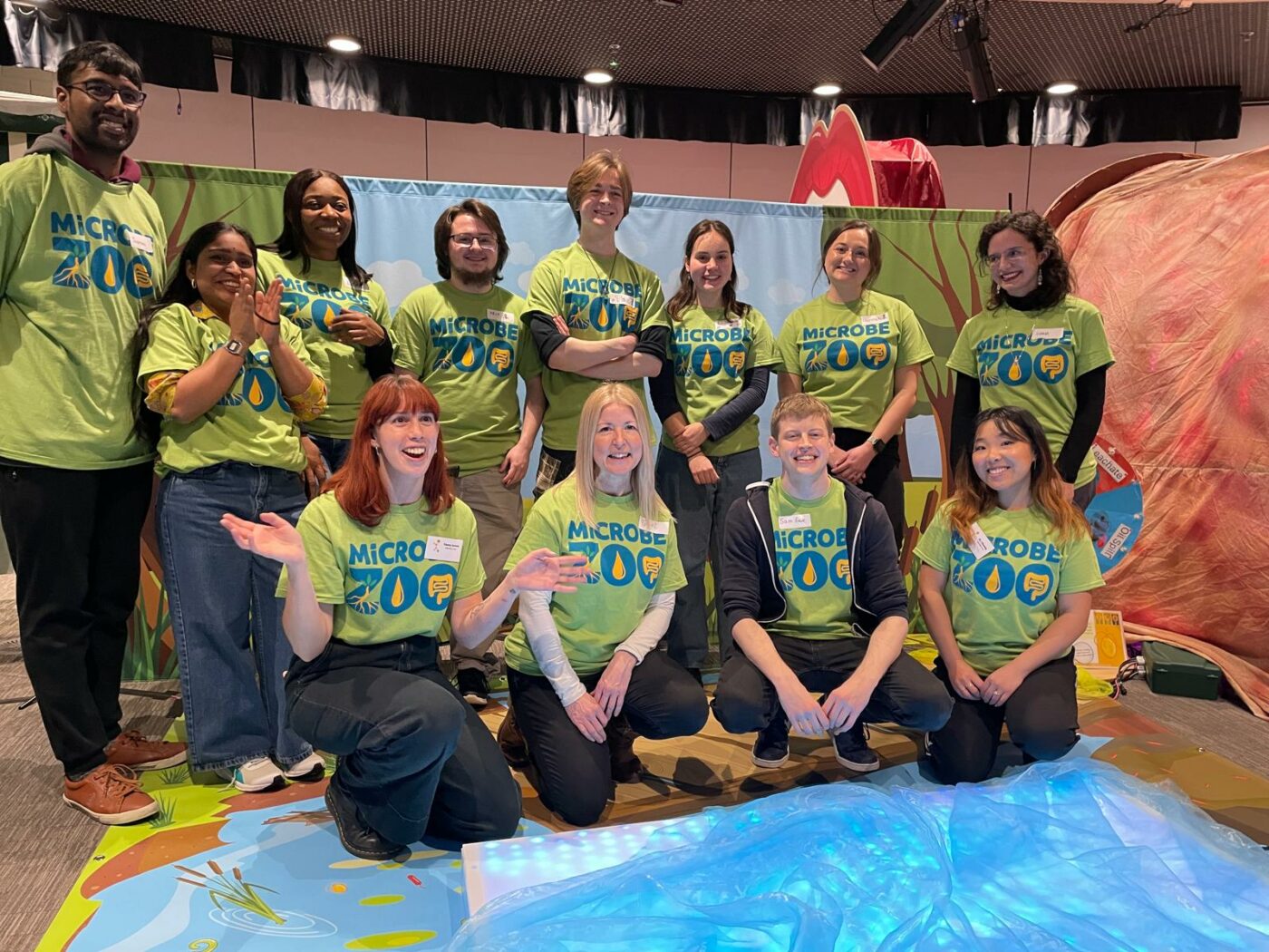 The Microbe Zoo team in matching lime-green shirts poses together at an indoor event, smiling at the camera with educational displays behind them.