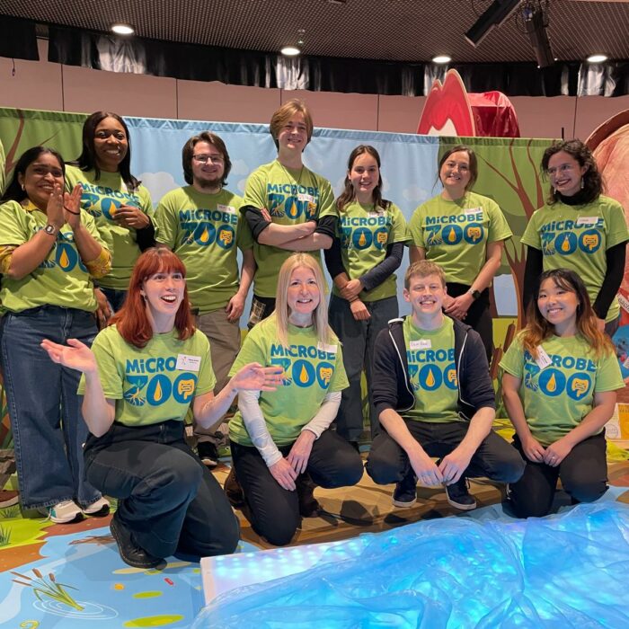The Microbe Zoo team in matching lime-green shirts poses together at an indoor event, smiling at the camera with educational displays behind them.